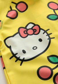 Fényes felületű sárga anyagon nagy Hello Kitty arc és cseresznye motívumok találhatók. A mintázat piros masnit és fekete körvonalakat tartalmaz.