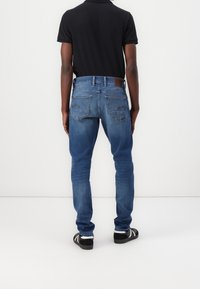 Jeans de mezclilla azul con corte ajustado, bolsillos traseros estándar y una etiqueta de cuero en la cinturilla. Llevados con zapatos negros y blancos.