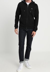 Sudadera negra con cremallera y cordones ajustables, bolsillos frontales y un pequeño logotipo. Combinada con vaqueros oscuros y zapatillas altas blancas.