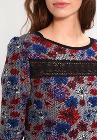 Gebreide top met bloemenpatroon, zwarte kanten accenten en een ronde halslijn. Heeft een mix van rode, blauwe en roze bloemen op een grijze achtergrond.