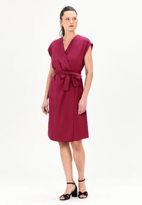 Vestido envolvente burdeos con mangas cortas, un cinturón ceñido en la cintura y un dobladillo a la altura de la rodilla, hecho de una tela suave. Combinado con tacones negros de tiras.