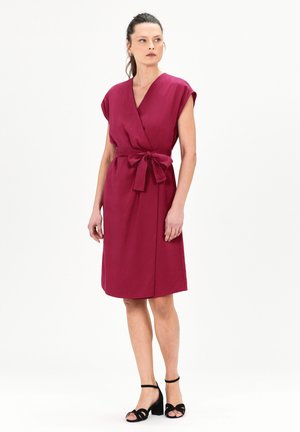 Bordeauxfarbenes Wickelkleid mit kurzen Ärmeln, einem gebundenen Taillengurt und einem knielangen Saum, gefertigt aus einem glatten Stoff. Kombiniert mit schwarzen Riemchensandaletten.