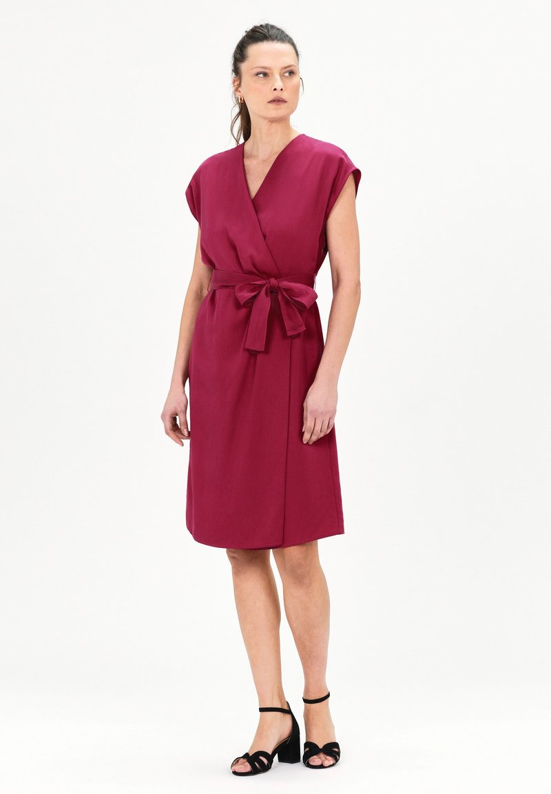 Vestido envolvente burdeos con mangas cortas, un cinturón ceñido en la cintura y un dobladillo a la altura de la rodilla, hecho de una tela suave. Combinado con tacones negros de tiras.
