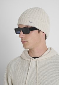Gorro de punto crema con textura acanalada y una pequeña etiqueta metálica, combinado con gafas de sol rectangulares negras con lentes oscuros. Usado con una sudadera ligera.