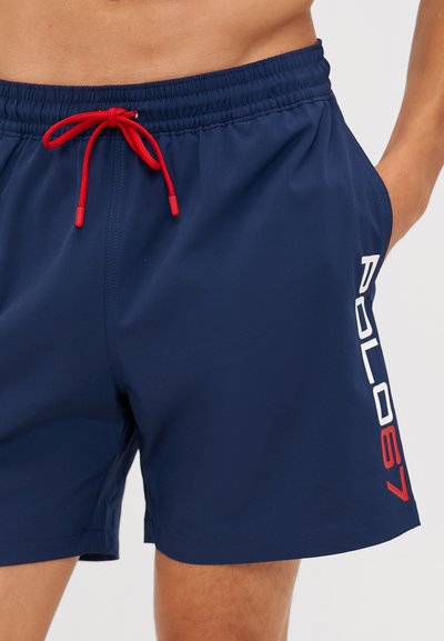 Shorts de bain bleu marine avec une taille élastique, un cordon rouge et des détails de logo blanc sur le côté. Tissu lisse et léger.