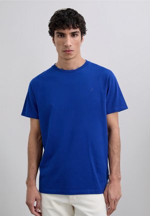 Young man with short curly hair wearing a plain blue short-sleeve t-shirt and white pants against a light gray background.