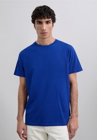 Giovane con capelli corti e ricci, indossa una maglietta blu a maniche corte e pantaloni bianchi su uno sfondo grigio chiaro.