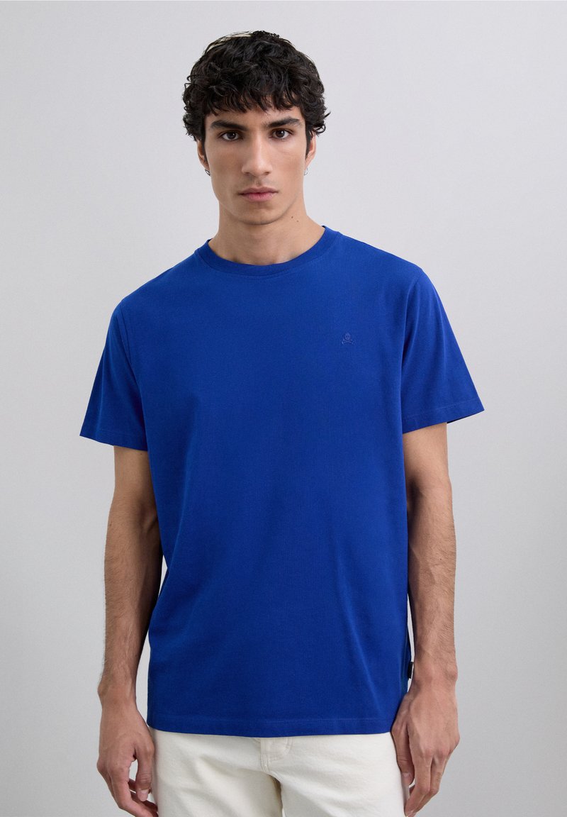Giovane con capelli corti e ricci, indossa una maglietta blu a maniche corte e pantaloni bianchi su uno sfondo grigio chiaro.
