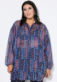 Mujer vestida con una blusa de botón de manga larga con un patrón geométrico y floral en azul, rojo y blanco, de pie frente a un fondo blanco.