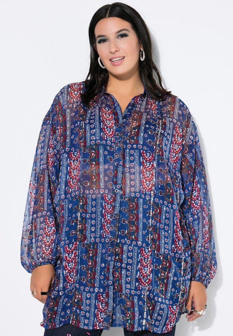 Mujer vestida con una blusa de botón de manga larga con un patrón geométrico y floral en azul, rojo y blanco, de pie frente a un fondo blanco.