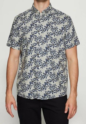 Mand iført en kortærmet skjorte med grå base og mørkeblå blomsterprint, der står foran en ensfarvet baggrund.