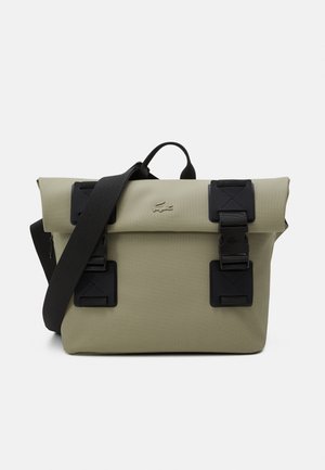 Oliwkowa torba na ramię z czarnym regulowanym paskiem, dwoma czarnymi klamrami oraz wytłoczonym logo krokodyla na przedniej klapce.