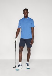 Μπλε μπλουζάκι πόλο με κοντά μανίκια, με γιακά και σχέδιο με ρίγα στο πλάι, σε συνδυασμό με σκούρα σορτς και λευκά παπούτσια για γκολφ. Κρατάει ρόπαλο γκολφ στο χέρι.