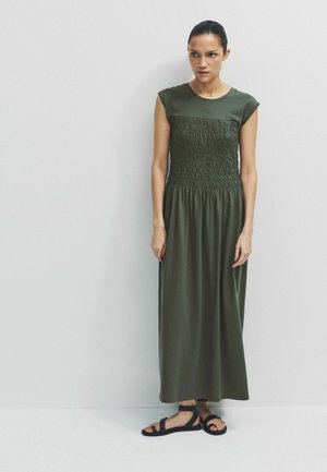 Femeie stând sprijinită de un perete, purtând o rochie maxi verde închis fără mâneci, cu bust strâns, și sandale negre plate.