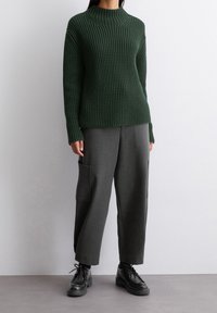 Zielony sweter dziany z wysokim kołnierzem, zestawiony z szerokimi szarymi spodniami z boczną kieszenią oraz czarnymi sznurowanymi butami.