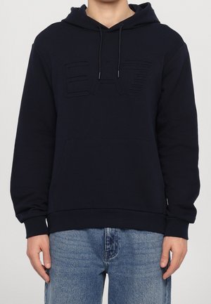 Mand iført en sort hættetrøje med "EA7" præget på brystet og blå jeans, stående mod en ensfarvet baggrund.
