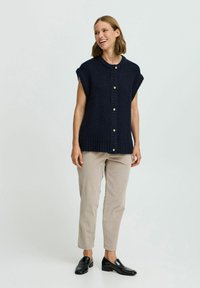 Marineblauer gestrickter Weste mit kurzen Ärmeln, Knopfschluss und geripptem Saum; kombiniert mit hellbeigen, schmalen Hosen und schwarzen Slippern.