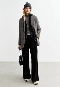 Houndstooth patroon jas met een zwarte fleece voering, grijze coltrui, zwarte wijde broek en crème sneakers. Zwarte tas in de hand.
