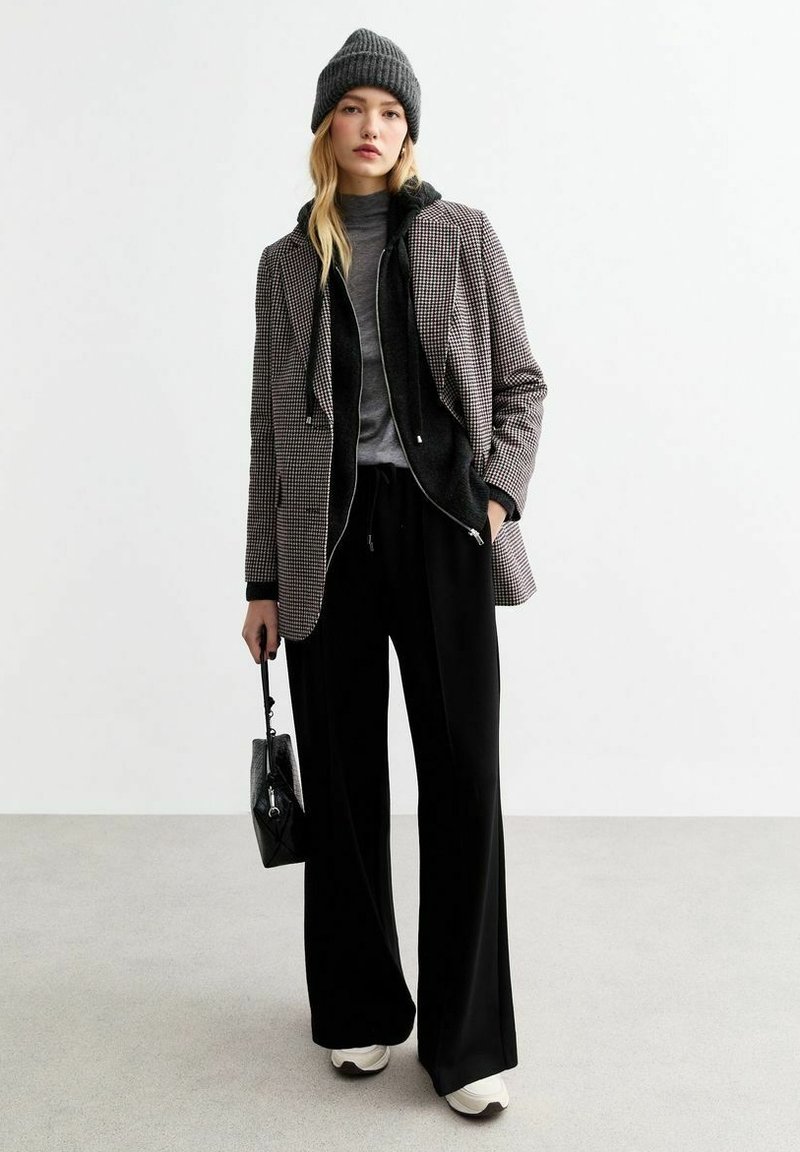 Giacca a motivo pied-de-poule con fodera in pile nera, dolcevita grigio, pantaloni neri a gamba larga e sneakers crema. Borsa nera in mano.