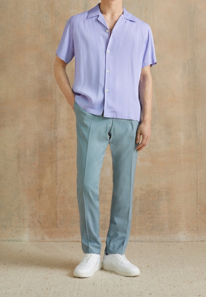 Camicetta corta lavender a maniche corte con bottoni, abbinata a pantaloni azzurri sagomati e sneakers bianche, su uno sfondo neutro.
