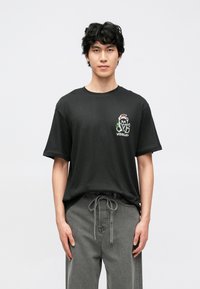 Tricou negru din bumbac, cu guler rotund, mâneci scurte și un design grafic pe piept care înfățișează un urs și cuvântul „VISIONARY”.