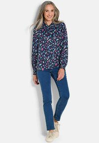 Marineblauwe blouse met lange mouwen en kleurrijke abstracte bladmotieven, gerimpelde halslijn, elastische boorden, gecombineerd met blauwe jeans en lichte sneakers.