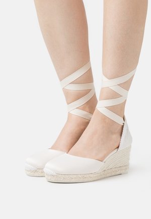 Des espadrilles blanches à compensés avec une tige en tissu, une semelle en jute tressé et des lanières croisées autour de la cheville. Texture lisse avec une subtile brillance.