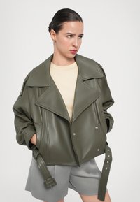 Veste en simili cuir vert olive avec un col oversize, quincaillerie argentée et détails de ceinture. Portée sur un haut beige clair et un short gris.