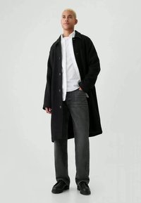 Μαύρο παλτό από μαλλί, με μεγάλα κουμπιά, φορεμένο πάνω από ένα λευκό πουκάμισο. Συνδυάζεται με γκρι τζιν και μαύρα παπούτσια, στέκεται σε απλό φόντο.