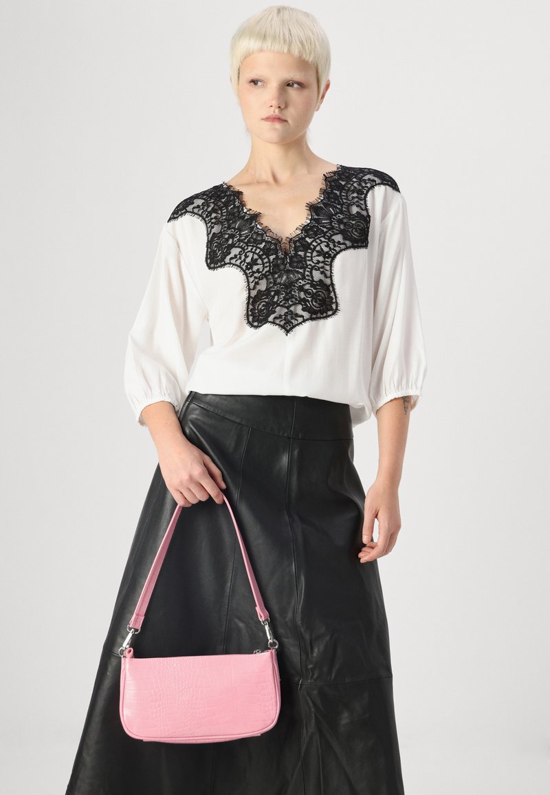 Chemisier blanc avec des détails en dentelle noire au niveau du col, associé à une jupe en cuir noir et un sac à main en cuir texturé crocodile rose clair.