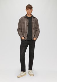 Jonge man die met zijn handen in zijn zakken staat, gekleed in een bruin geruite blouse, zwart t-shirt, zwarte spijkerbroek en beige sneakers.