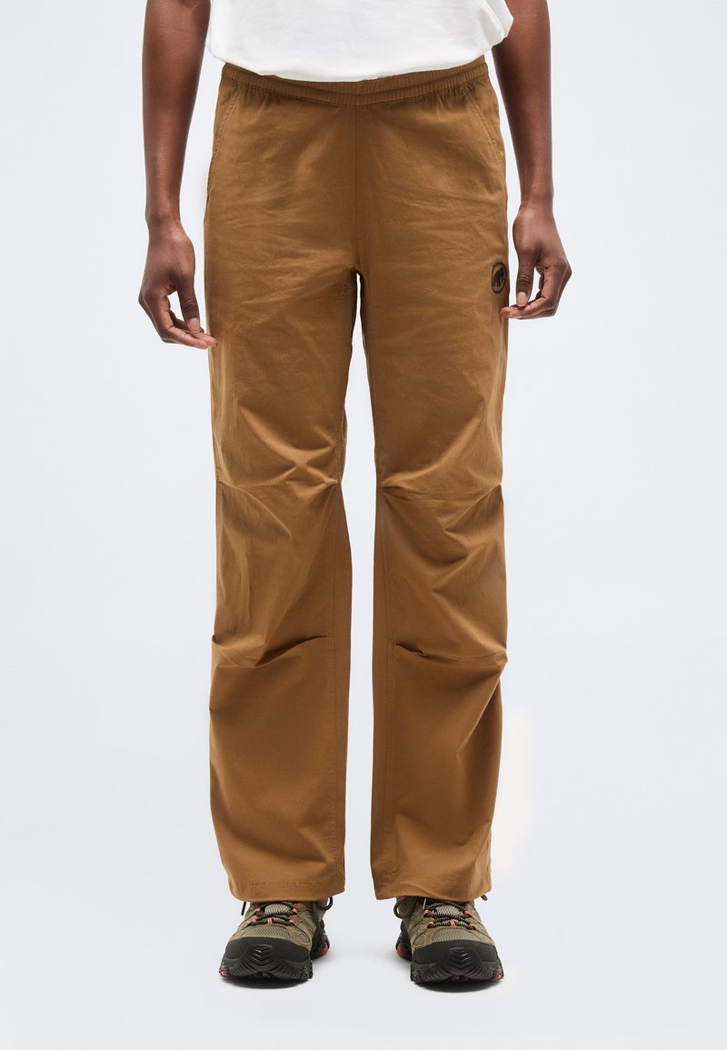 Άτομο που φοράει καφέ παντελόνια με ίσια γραμμή και παπούτσια πεζοπορίας, στέκεται μπροστά σε απλό φωτεινό φόντο.