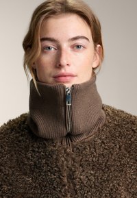 Bruine fleecejas met een hoge geribbelde kraag en zilveren ritsdetail. De textuur is zacht en pluizig, en biedt warmte en comfort.