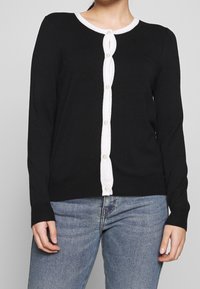 Svart långärmad kofta med vit knapplist och kant. Tillverkad av mjukt stickat material, med rund halsringning och ribbade ärmmuddar.