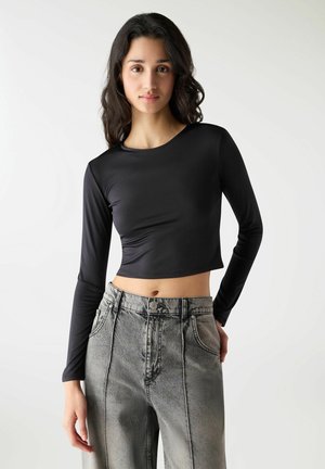 Młoda kobieta z długimi ciemnymi włosami, ubrana w czarny, długi top odsłaniający brzuch i szare jeansy z wysokim stanem, stojąca na białym tle.