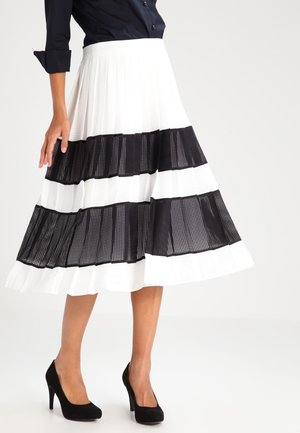 Vrouw draagt een witte geplooide midi-rok met twee brede zwarte horizontale mesh strepen, gecombineerd met zwarte hoge hakken en een donker topje.