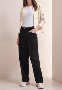 Chaqueta bomber crema con un patrón intrincado, camiseta blanca, pantalones anchos negros y zapatillas blancas, de pie contra un fondo neutro.