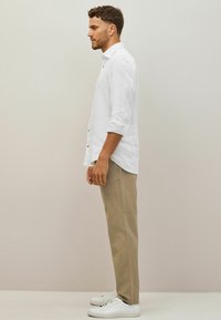 Vit långärmad linneskjorta med knappar, beige chinos och vita sneakers, sedd från sidan mot en neutral bakgrund.
