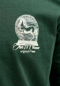 Έμβλημα λευκού αλόγου και κείμενο "The IVY Manor originals 1990" τυπωμένα σε σκουρό πράσινο ύφασμα.