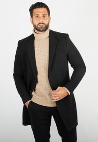 Manteau noir à revers étroits porté par-dessus un pull en laine torsadée beige à col montant. Un pantalon noir complète le look. Fond neutre.