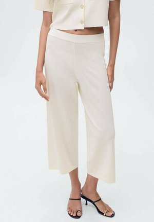 Pantalons larges crème avec une texture lisse, dotés d'une taille élastique et d'une coupe raccourcie, associés à des sandales noires à lanières.