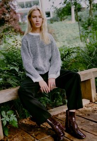 Hallikas ja valge triibuline kudumipusa laia mansjetiga, kombineeritud mustade pükstega ja läikivate burgundi värvi ankle boot'idega. Loomulik taust.