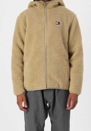 Béžová fleecová bunda na zip s kapucí, texturovaná, jemná látka a malé logo Tommy Jeans na levé straně hrudi, nošená s šedými kalhotami.