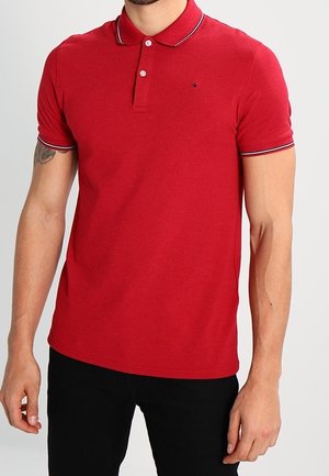 Mand iført en rød kortærmet poloshirt med hvide og sorte kanter på kraven og ærmerne, kombineret med sorte bukser.