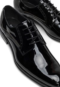 Zapatos de vestir de charol negro con punta afilada, diseño de cordones y perforaciones decorativas a lo largo de la parte superior. Acabado liso y brillante.