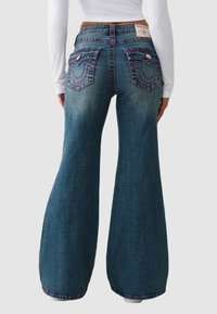 Dżinsy dzwony z ciemnoniebieskiego denimu z charakterystycznymi zakrzywionymi tylnymi kieszeniami, różowymi przeszyciami i wysokim stanem.