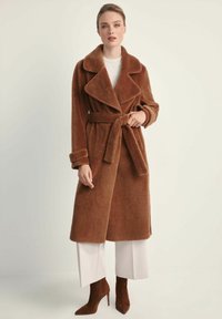 Kobieta w brązowym futrzanym płaszczu z szerokim paskiem, białych szerokich spodniach i brązowych wysokich butach na obcasie, stojąca na tle jednolitego jasnego tła.