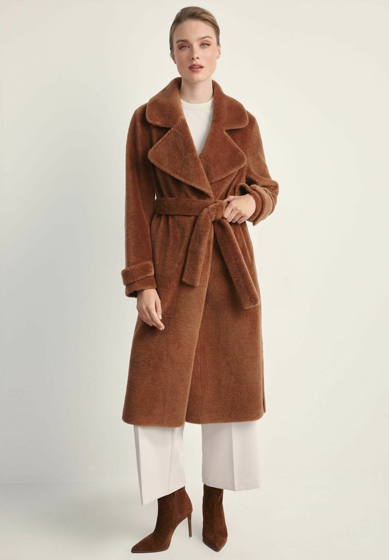 Kobieta w brązowym futrzanym płaszczu z szerokim paskiem, białych szerokich spodniach i brązowych wysokich butach na obcasie, stojąca na tle jednolitego jasnego tła.