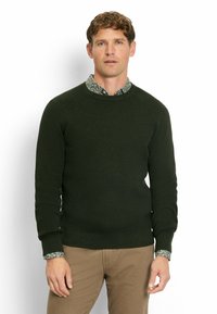 Dunkelgrüner Strickpullover mit gerippter Textur und Rundhalsauschnitt, über einem Hemd mit floralem Muster und mit Knopfmanschetten getragen.