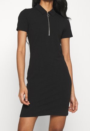 Žena v černých polokošilových šatech s krátkým rukávem, předním zipem a límečkem, stojící před jednobarevným bílým pozadím.